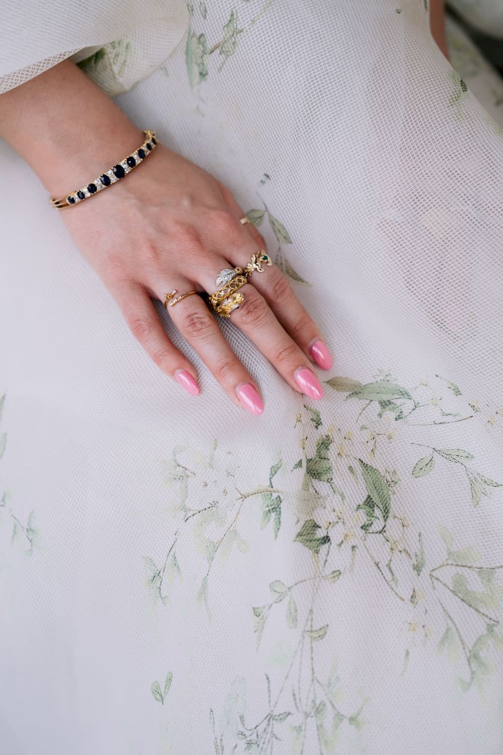 Bride's hand with vintage sapphire bracelet and stacked gold rings resting on floral-embroidered skirt.