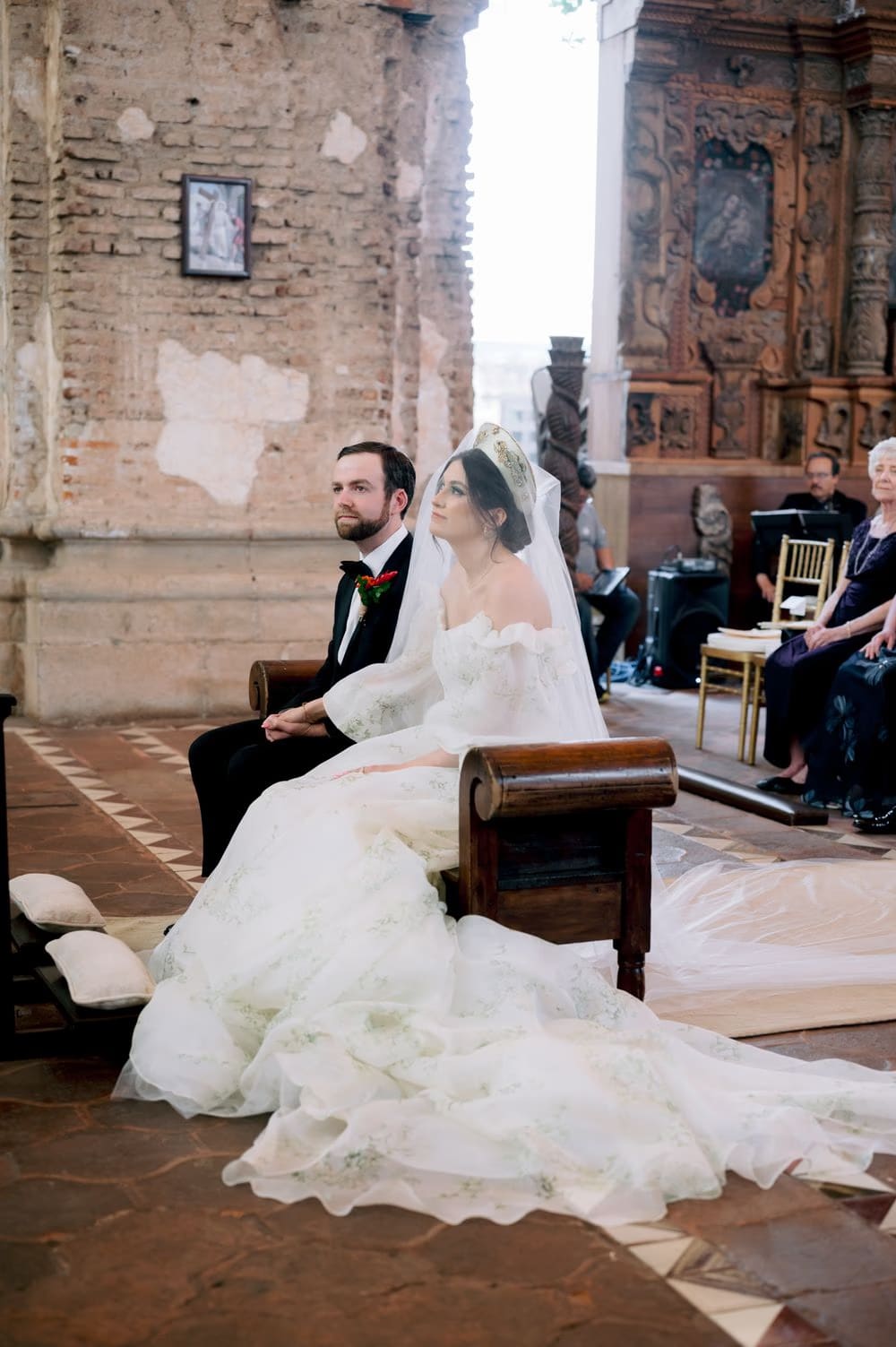 Bride and groom kneel together at a baroque altar with tropical florals during their wedding ceremony.