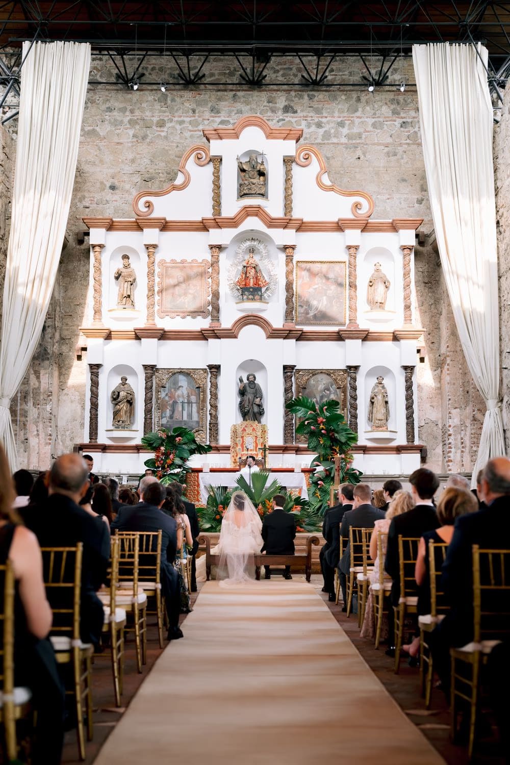 Wide ceremony shot showing white draped curtains, baroque altarpiece and tropical arrangements at Casa Santo Domingo chapel — destination wedding in Antigua Guatemala.