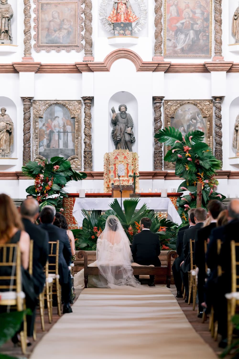 Bride sits before the baroque altar framed by tropical palm arrangements during the ceremony at Hotel Casa Santo Domingo, Antigua Guatemala.