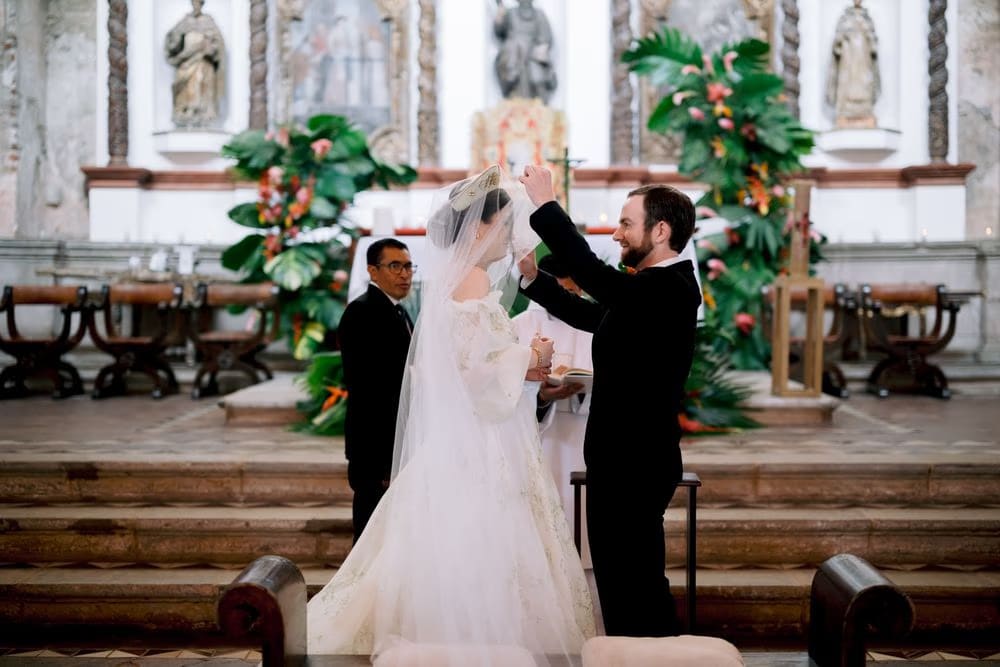Groom lifts the bride's veil over her crown during the unveiling ritual at the chapel altar.
