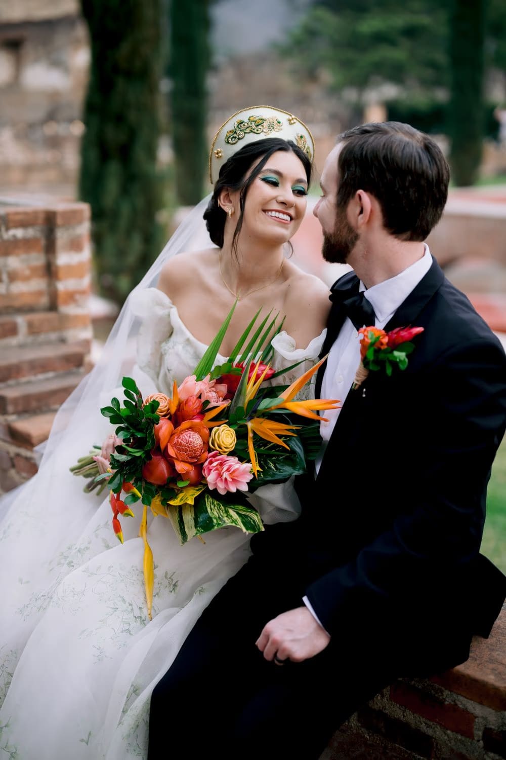 Bride and groom share a close look on a brick wall, tropical bouquet between them — golden-hour couple portrait in Antigua.