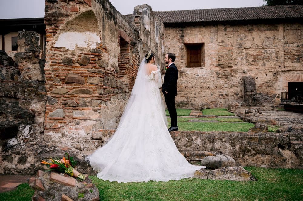 Bride and groom face each other inside a stone arch among monastery ruins — wide editorial couple portrait.