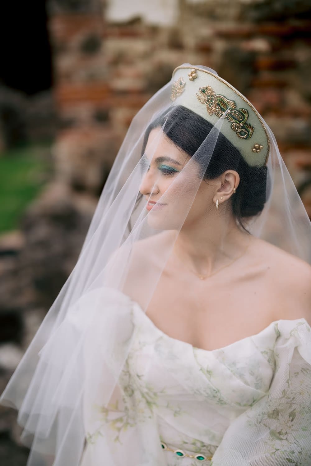 Bridal portrait in soft profile, veil draped over the shoulders against weathered brick — beaded crown with emerald motifs.