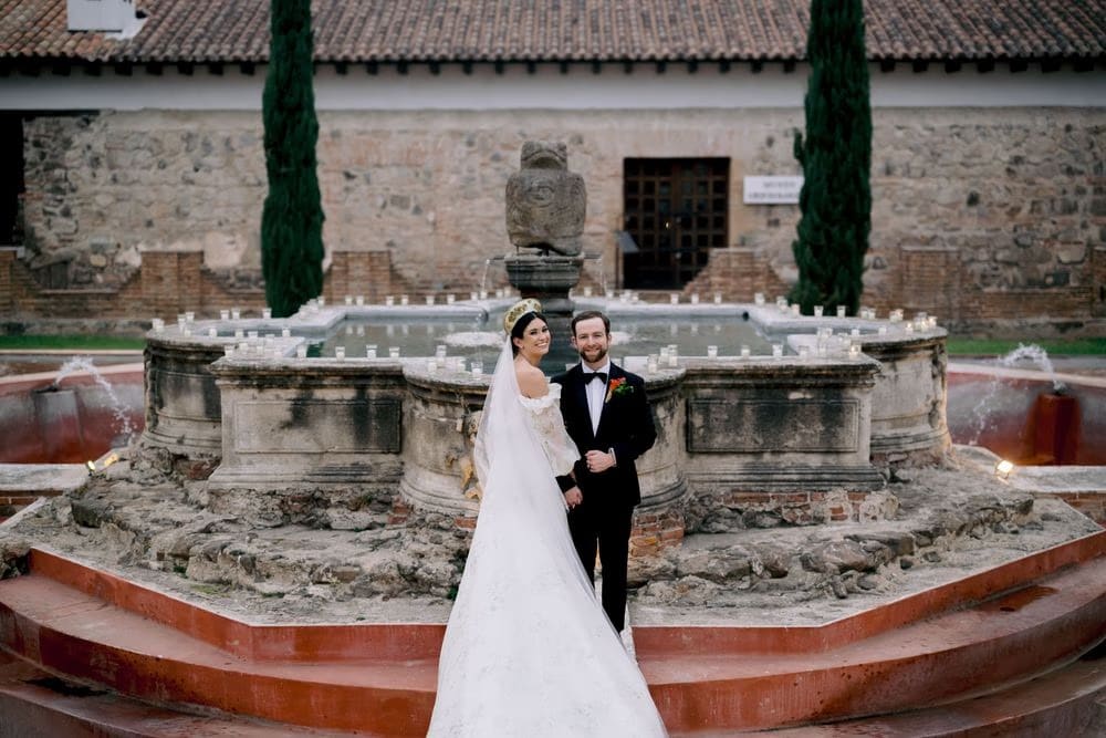 Newlyweds pose by the candle-lit central fountain at Hotel Casa Santo Domingo at dusk, Antigua Guatemala.