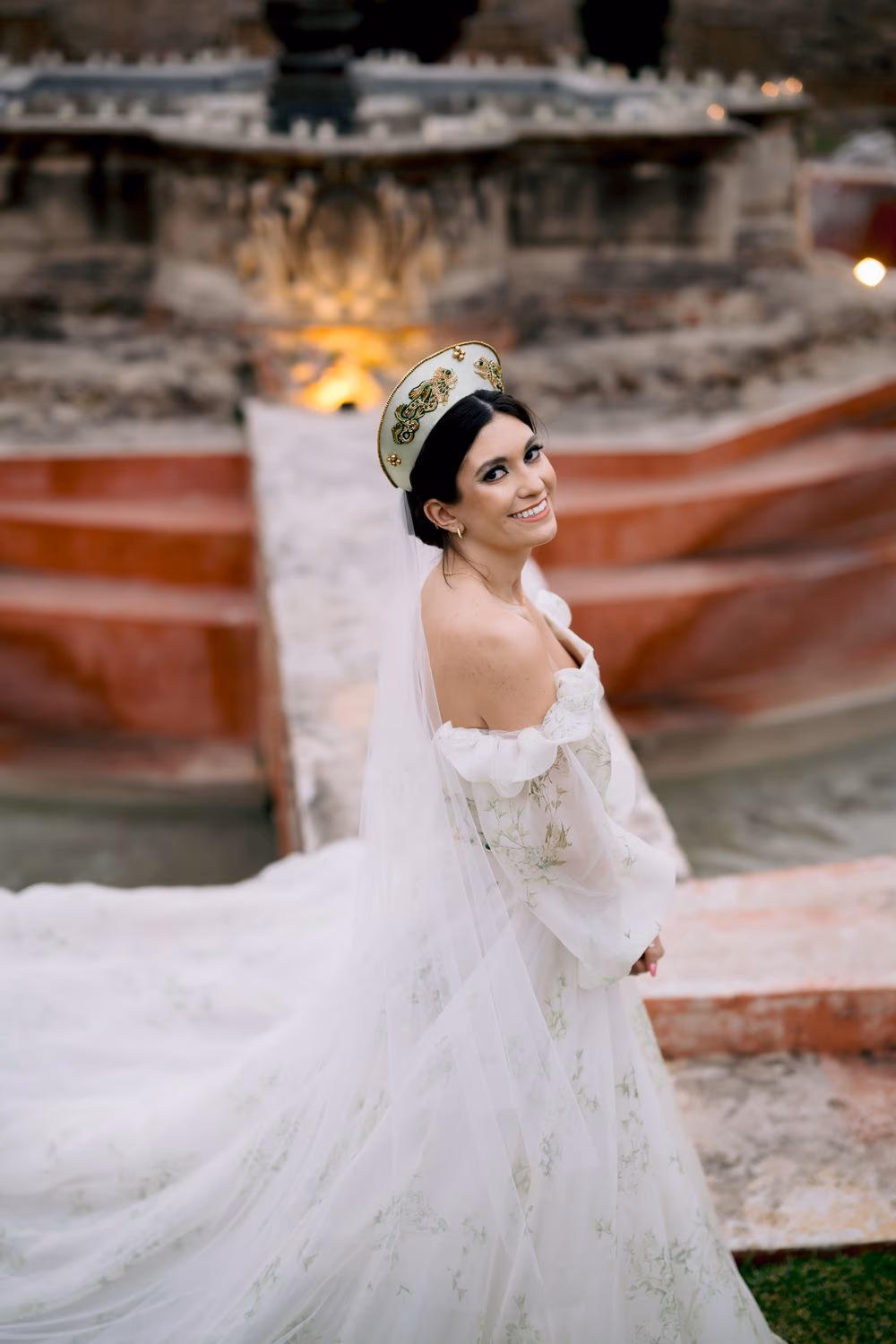 Bride smiles over her shoulder in front of a candle-lit fountain at dusk — golden-hour bridal portrait.