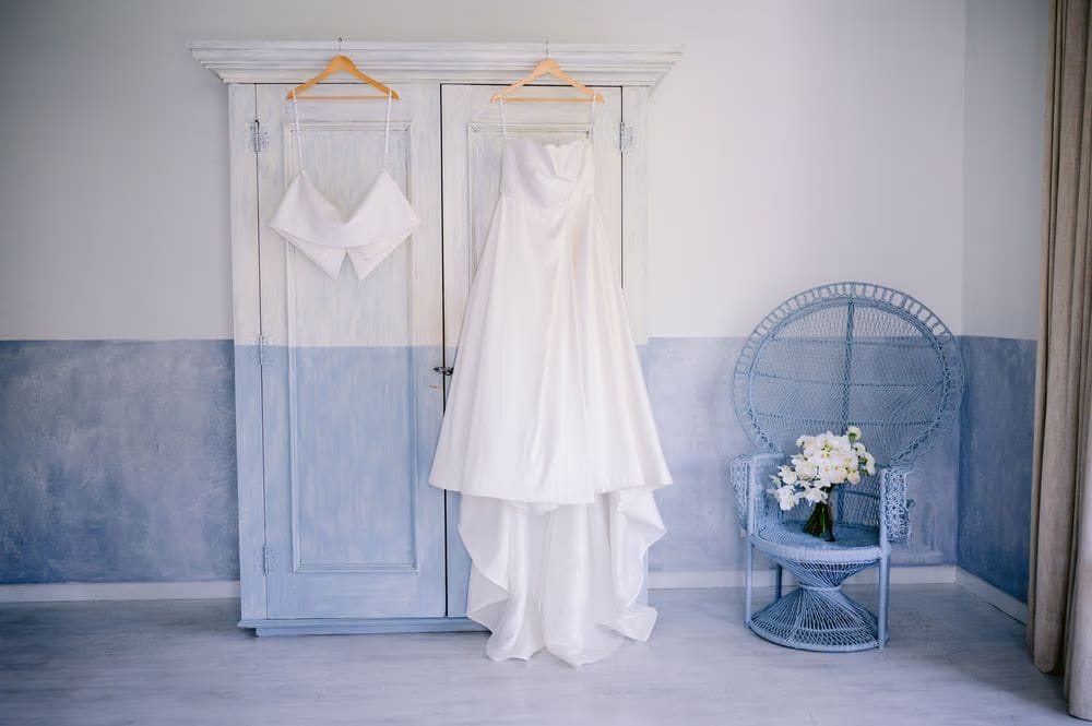 The bride's gown and stole hang from a whitewashed armoire beside a wicker peacock chair on the morning of the wedding at Villa Bokeh.