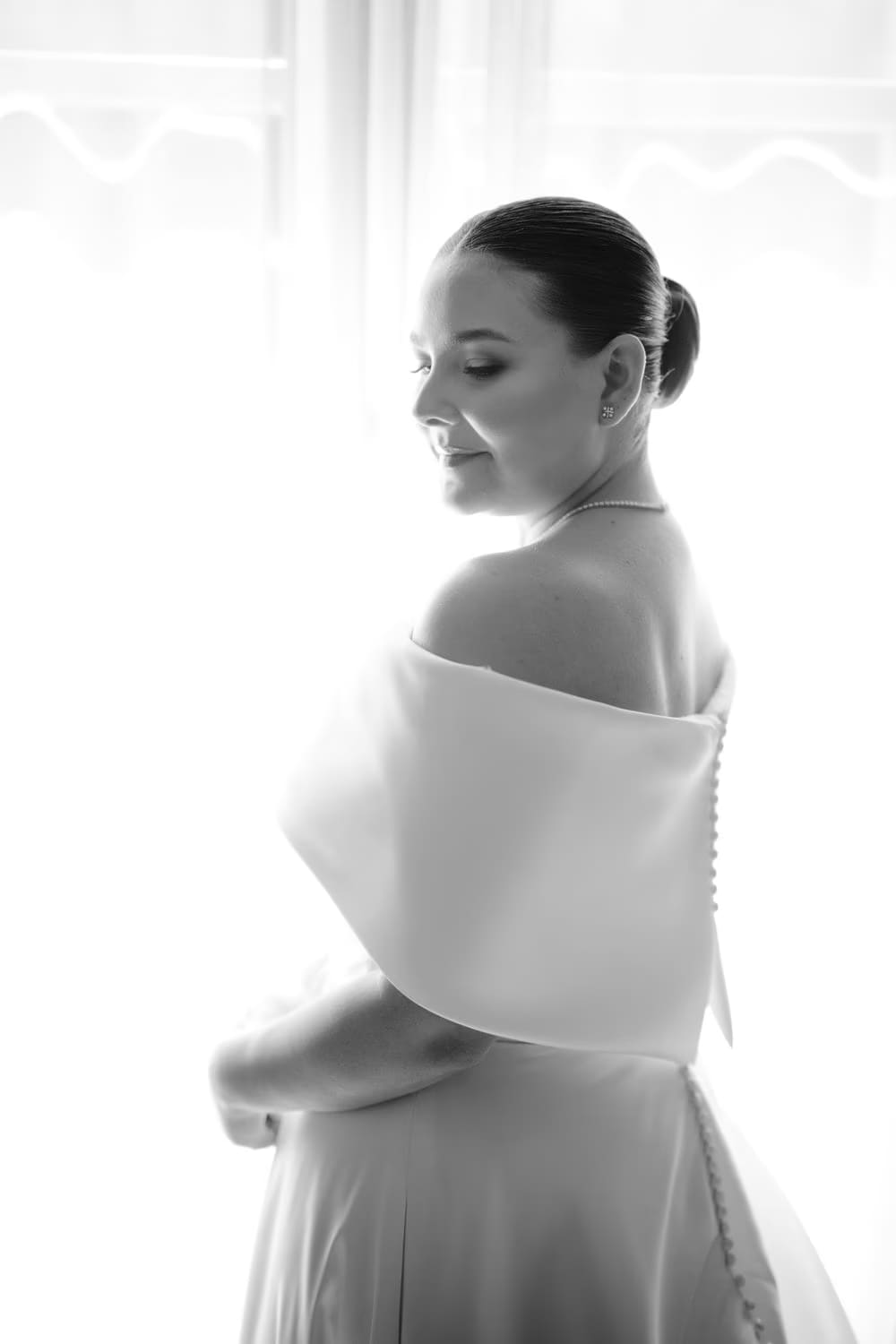 Black-and-white study of the bride from behind, light pouring through sheer curtains.