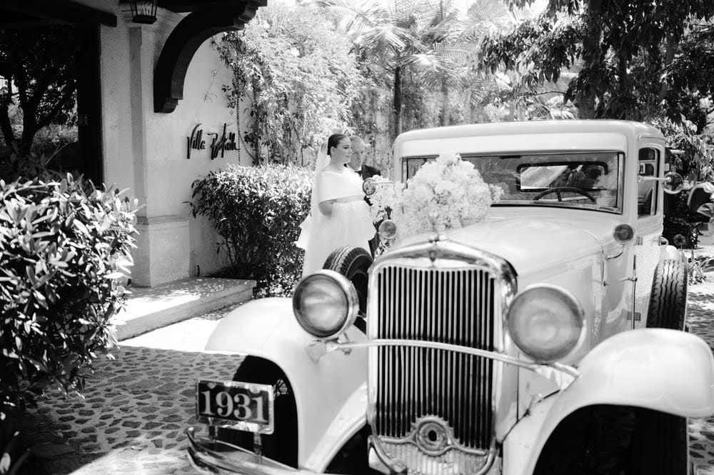 Bride arrives at the venue beside a vintage 1931 sedan dressed with white florals — black-and-white film aesthetic.