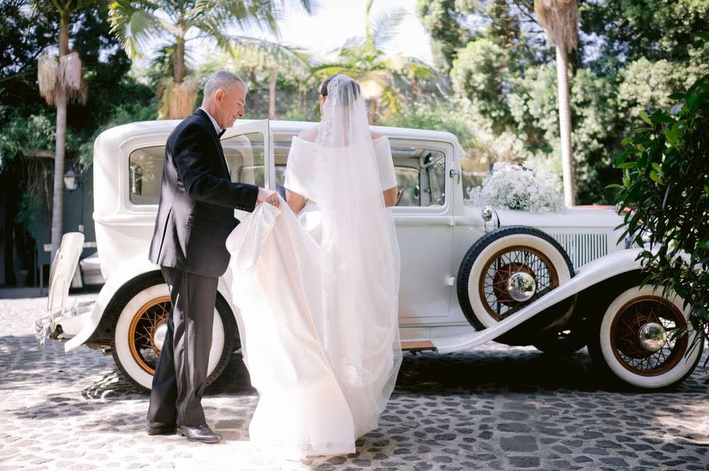 Father gathers Johanna's train as she steps toward the antique car decorated with baby's breath — Antigua Guatemala wedding.