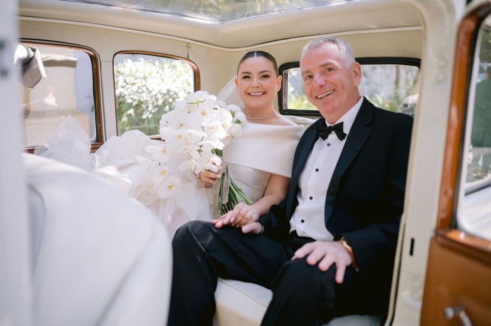 Bride and her father smile inside the vintage car with her orchid bouquet on the way to the ceremony.