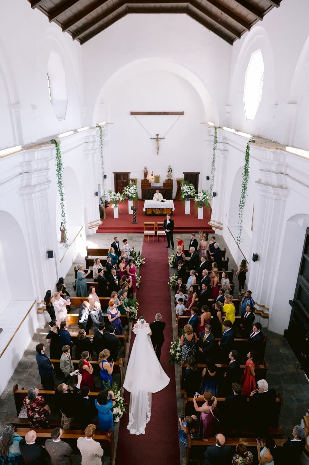 Aerial perspective of the bride walking down the long red carpet aisle of the colonial church.