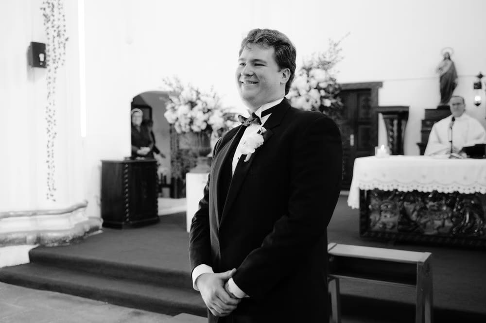 Groom Thomas smiles as he watches the bride approach the altar — black-and-white ceremony moment.