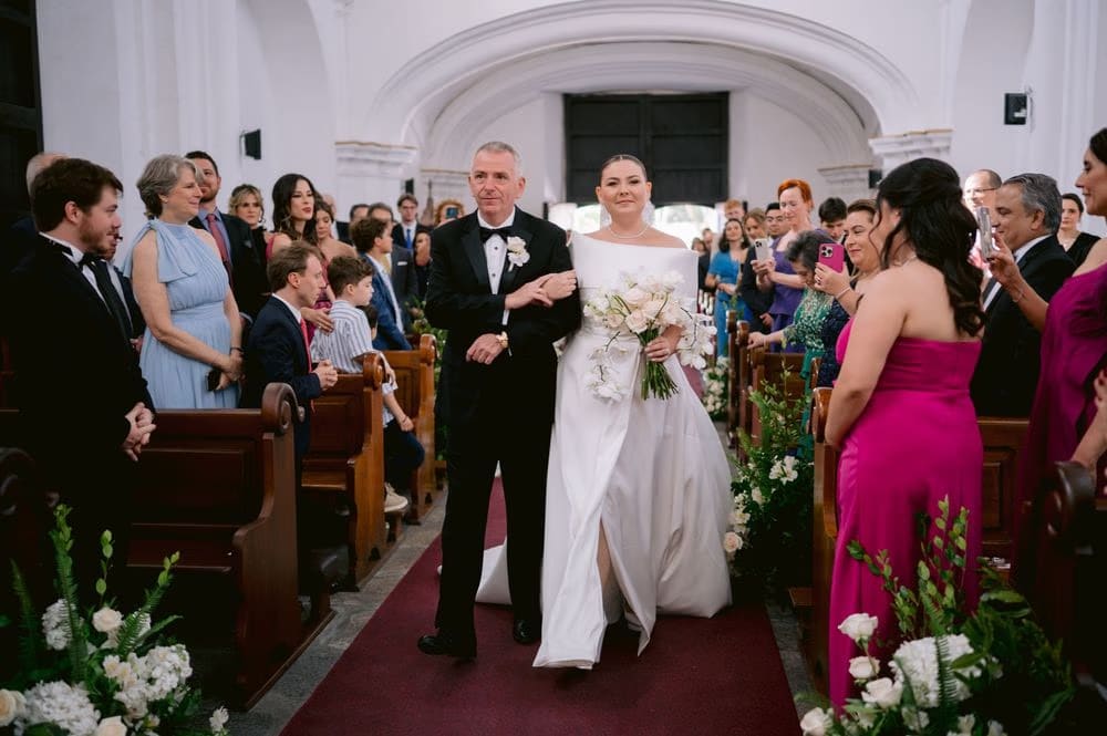 Johanna walks down the aisle on her father's arm holding a cascading white bouquet — wedding ceremony in Antigua Guatemala.