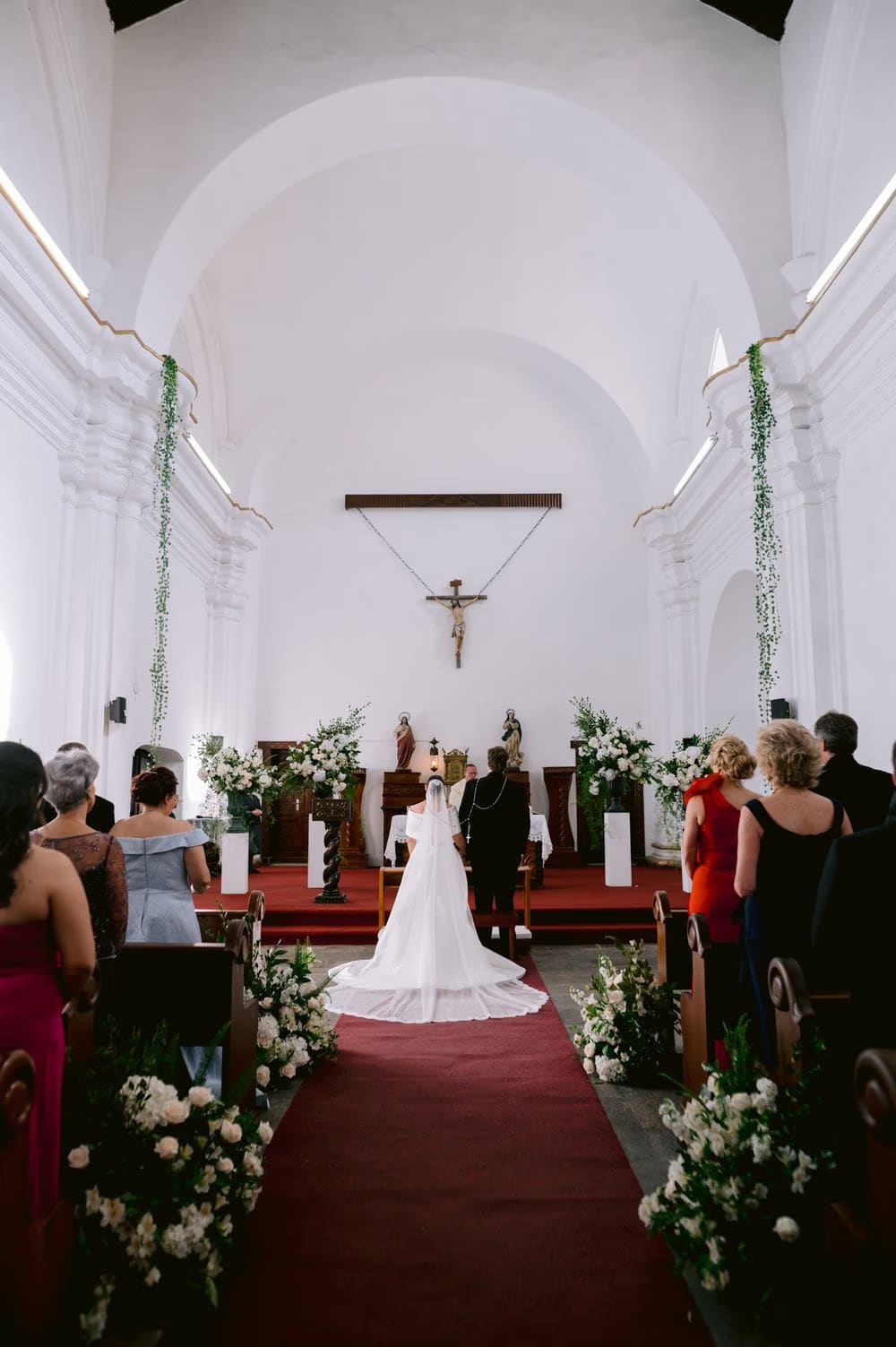 View from the aisle of Johanna at the altar before the crucifix, her cathedral train fanned across the red carpet.