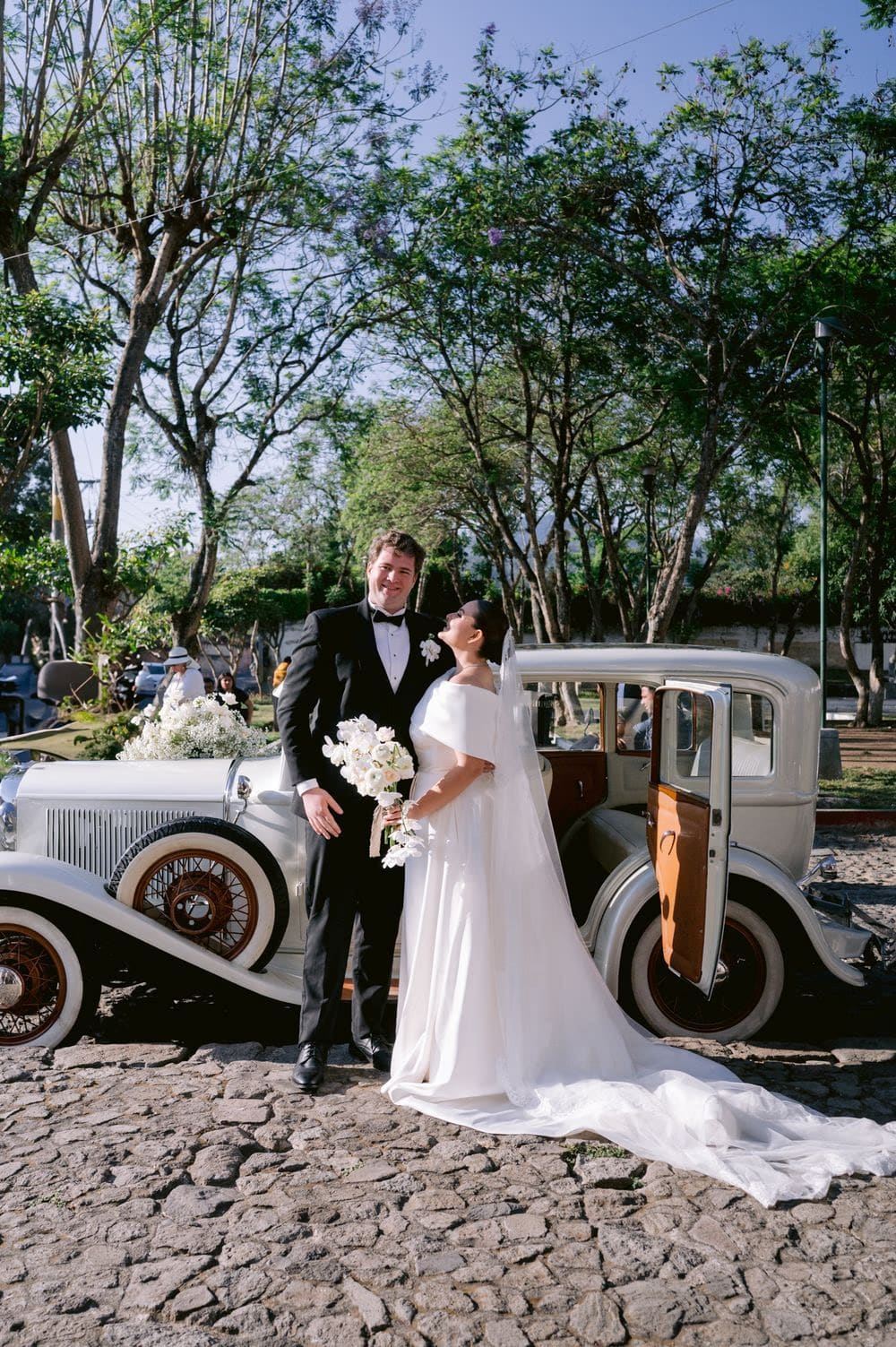 Bride and groom portrait beside the vintage white wedding car on a cobbled lane — Antigua Guatemala.