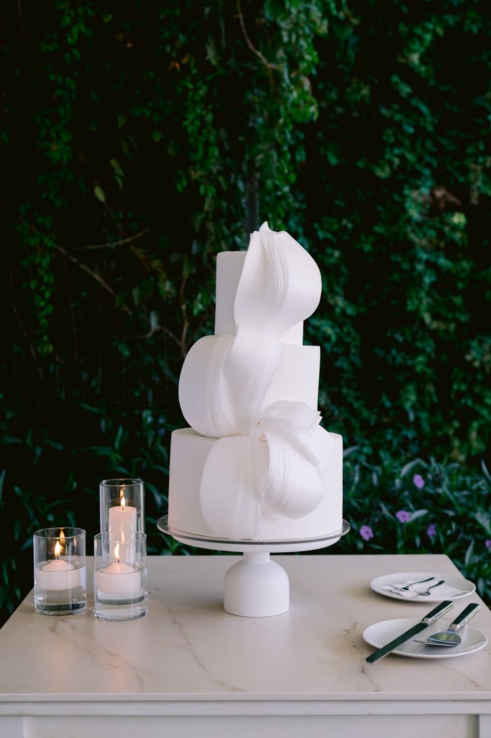 Three-tier sculptural white wedding cake with sail-shaped fondant ribbons against a green ivy backdrop.