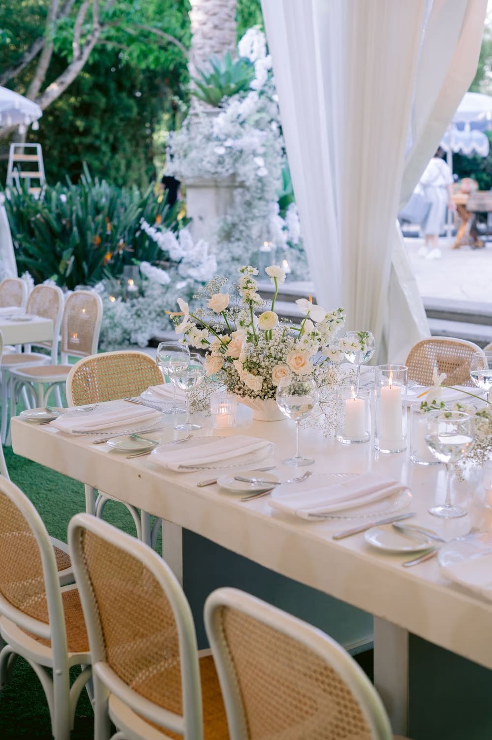 Long reception table dressed in cream linen with cane chairs, candles and a low white centerpiece.