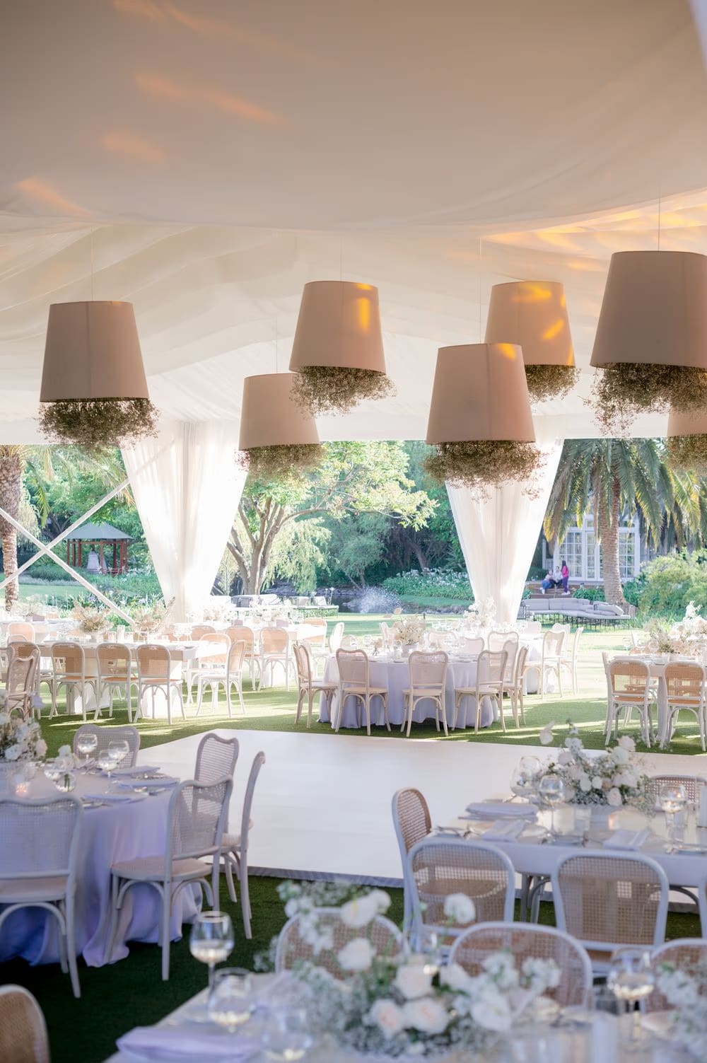 Reception tent at sunset with hanging lamp shades wrapped in baby's breath, round tables and a central dance floor.