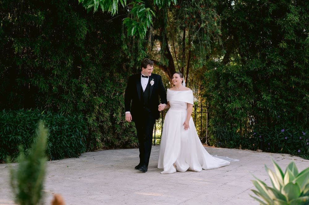 Bride and groom walk hand-in-hand through a leafy archway in the tropical gardens.