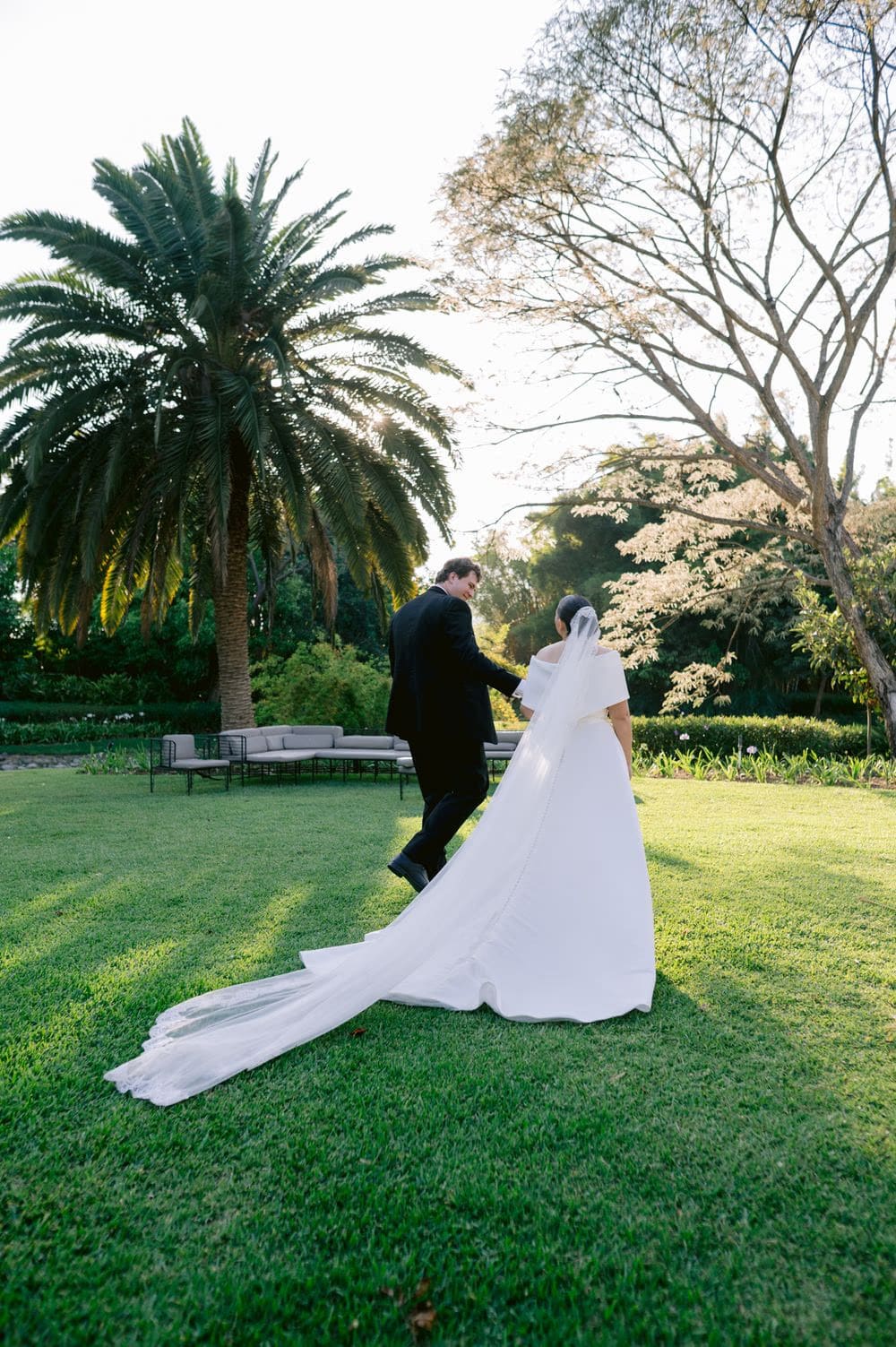 Bride and groom walk arm-in-arm across the lawn beside a tall palm — destination wedding at Villa Bokeh.