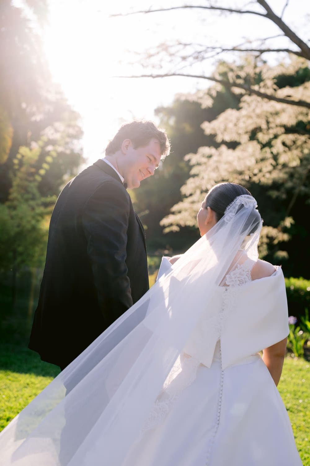 Sunlit portrait of the groom leading the bride by the hand through the gardens at golden hour.