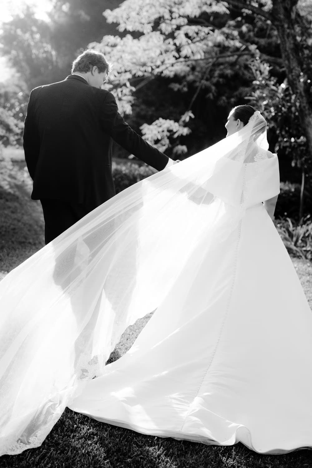 Wind catches the bride's cathedral veil and gown train as the couple walks together — editorial black-and-white portrait.