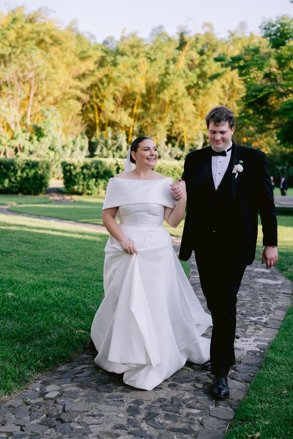 The bride lifts her train as the groom escorts her along a flagstone path through the gardens.