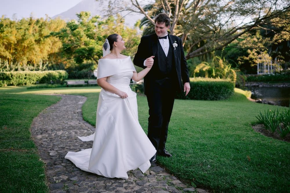 The groom twirls the bride along a cobblestone path bordered by neat hedges — playful wedding portrait.