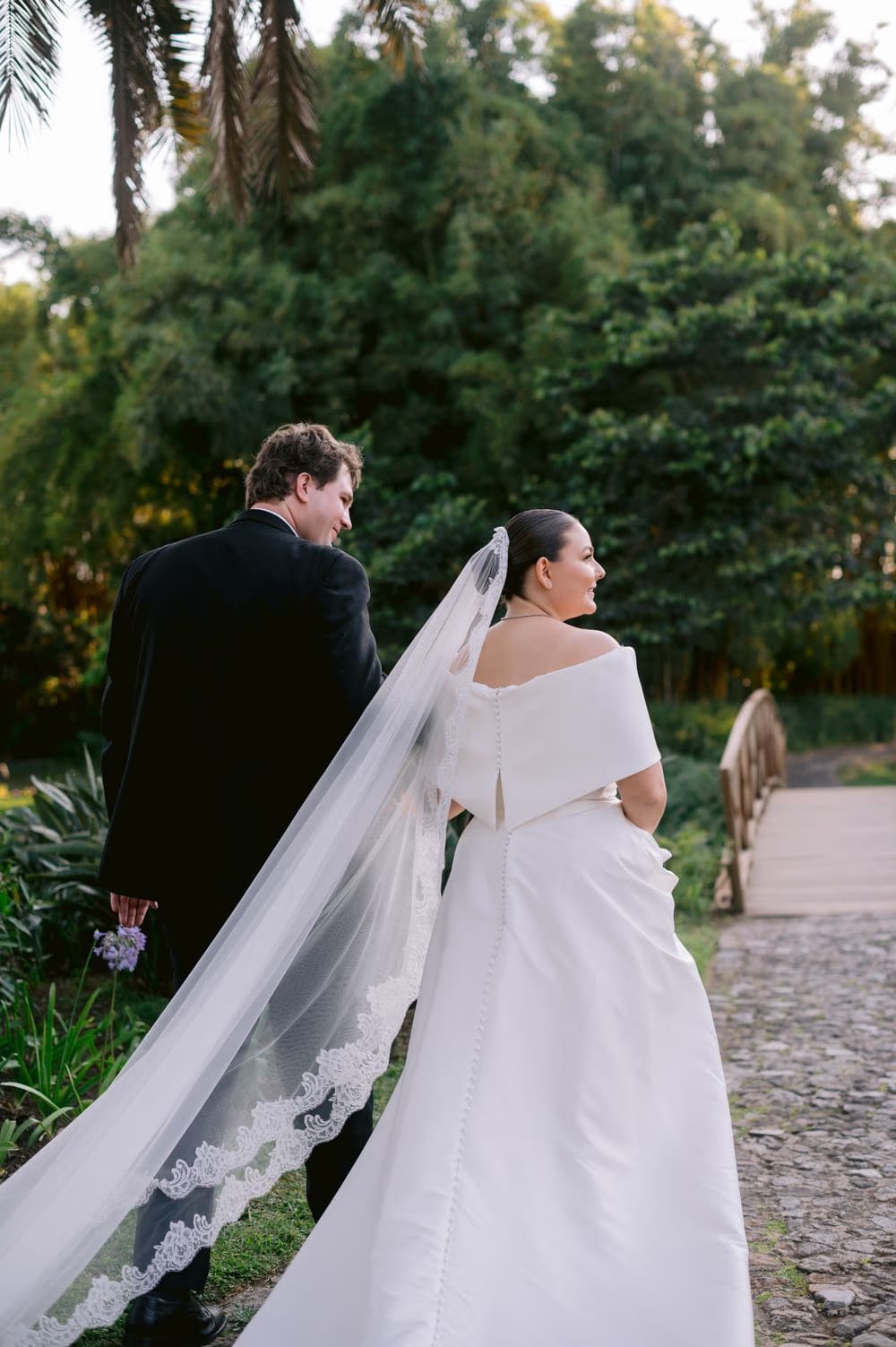 Bride and groom walk away across the wooden footbridge, her cathedral veil trailing behind them.