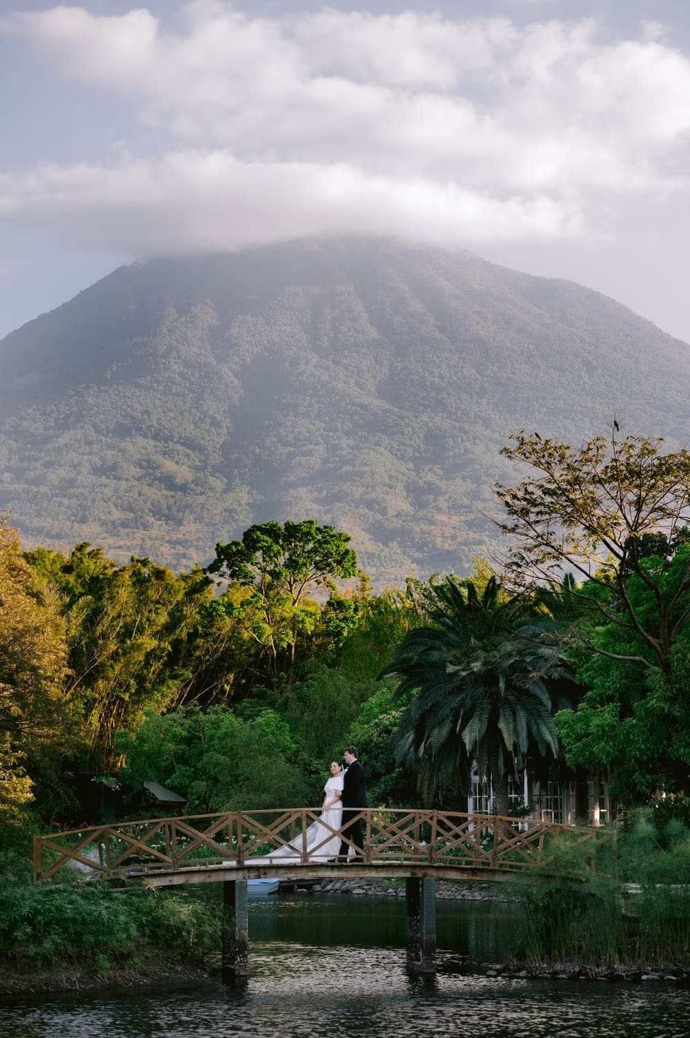Couple poses on the wooden bridge with the cloud-wrapped volcano towering behind — open-air portrait at Villa Bokeh.