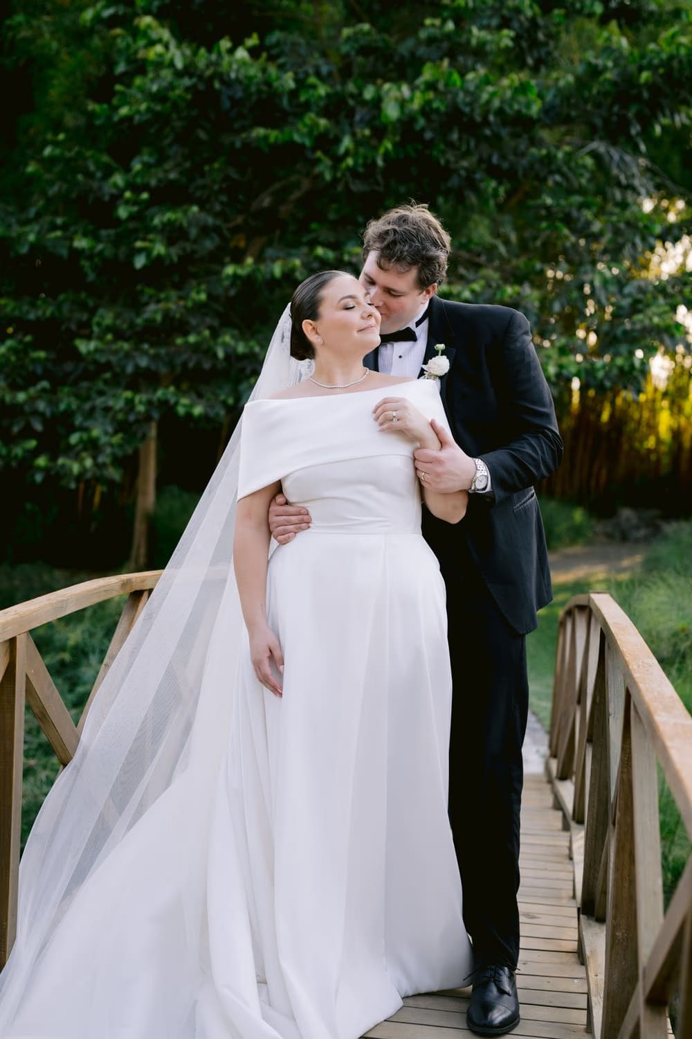Thomas kisses Johanna's forehead on the wooden bridge over the pond — intimate sunset moment at Villa Bokeh.