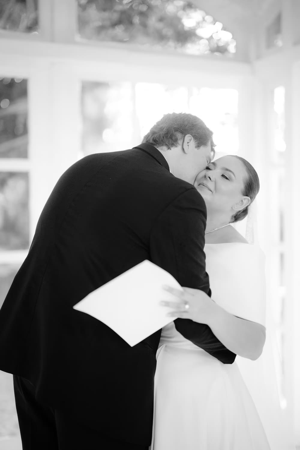 Black-and-white moment of the groom kissing the bride's cheek after sharing private vows.