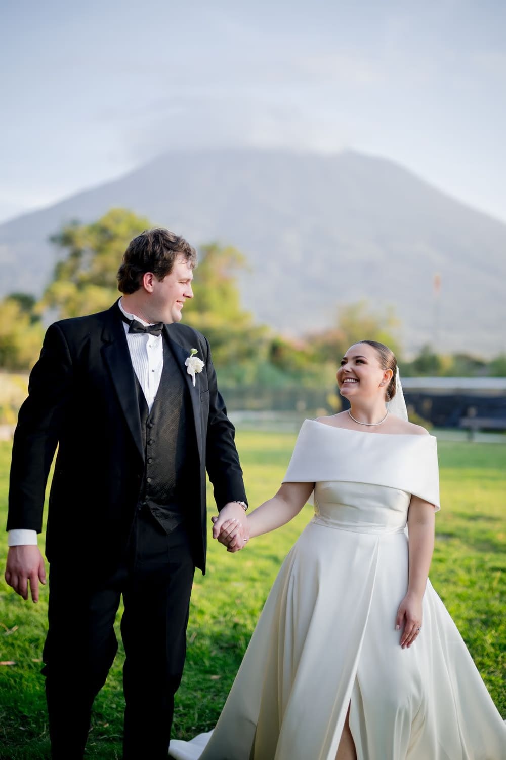Bride and groom hold hands on the lawn with the volcano shrouded in clouds behind them — Antigua Guatemala wedding.