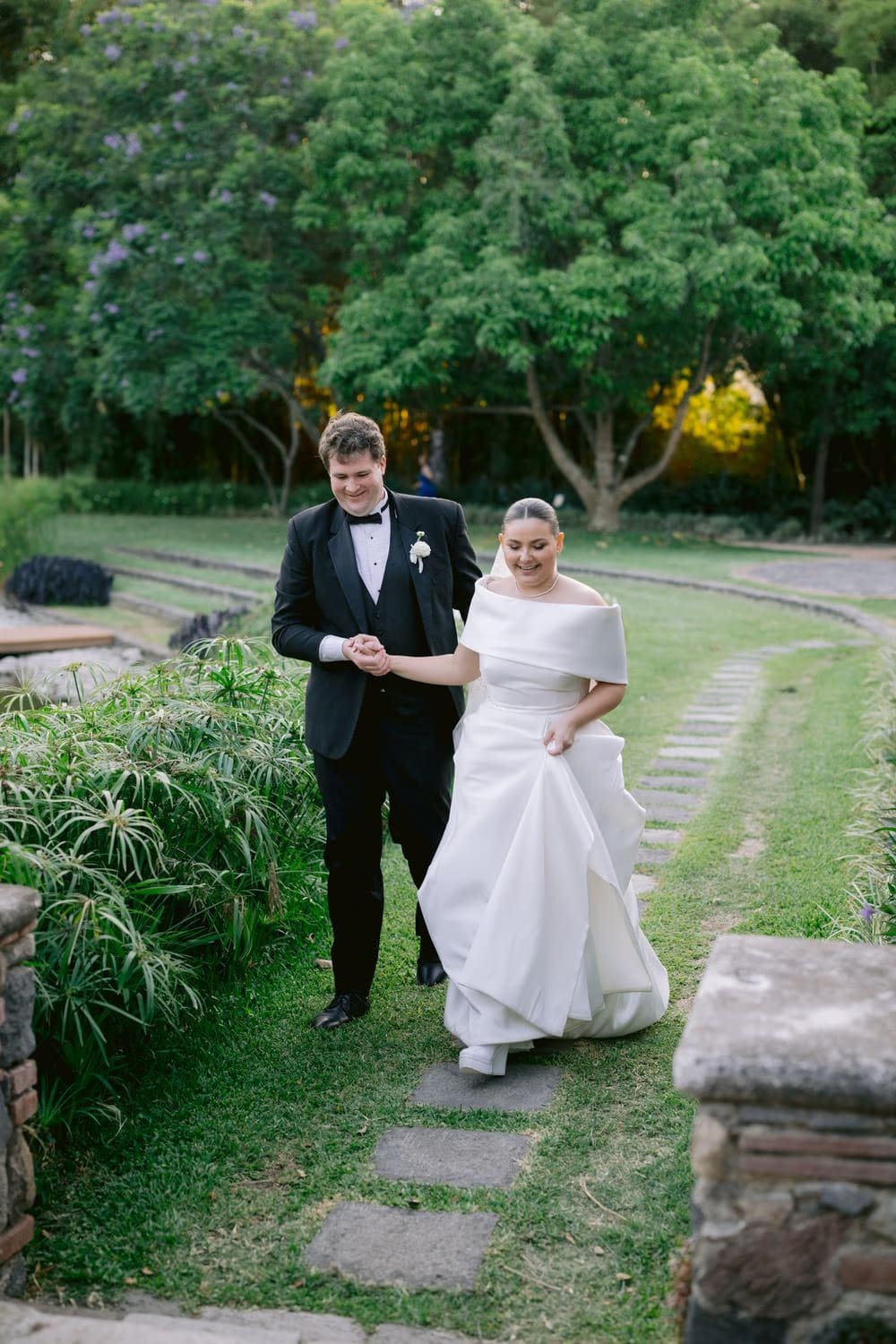 Couple walks a flagstone path lined with jacaranda blooms in the gardens at Villa Bokeh.