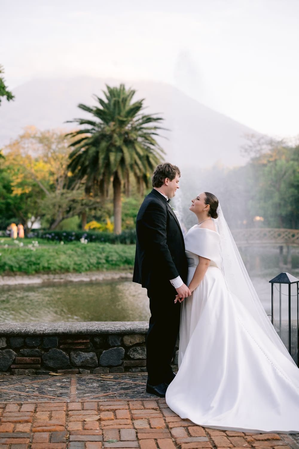 Bride and groom pause on a stone terrace with palms and a misty volcano framing them at dusk.