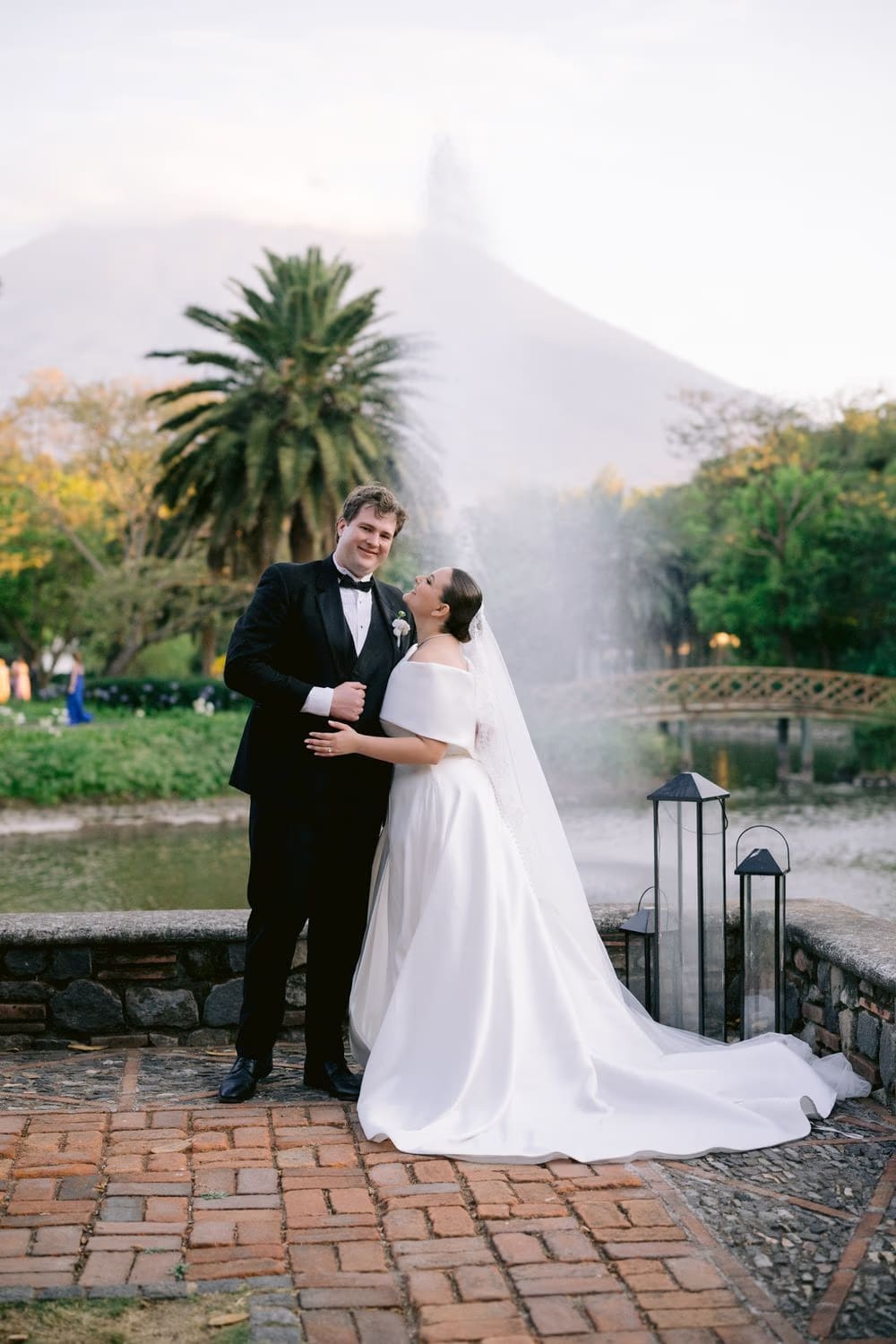 Newlyweds pose beside the pond with palm trees and the volcano emerging from mist behind them.