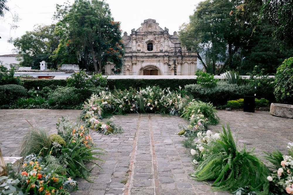 Outdoor ceremony aisle framed by ferns and wildflowers facing the San José el Viejo Ruins facade in Antigua Guatemala.