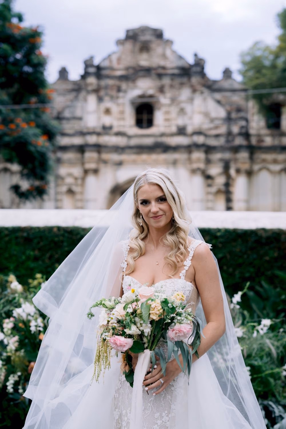 Bride Sydney holding a wild garden bouquet with veil flowing in front of the San José el Viejo facade — editorial bridal portrait in Antigua Guatemala.