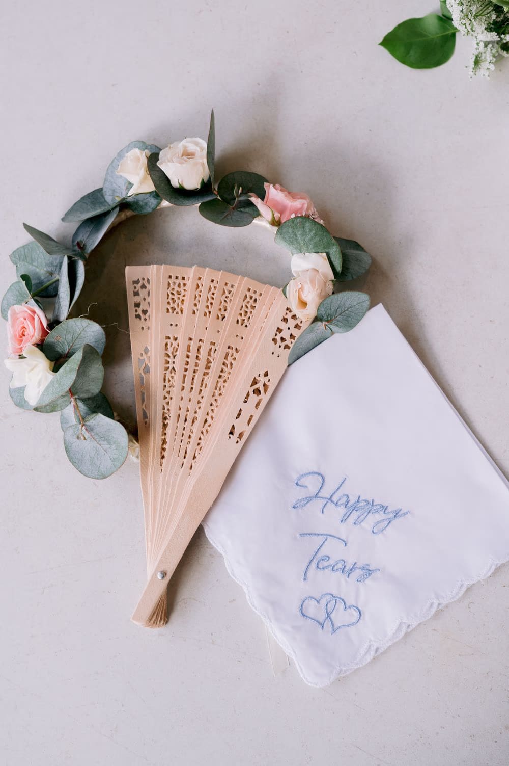 Wooden hand fan, eucalyptus flower crown and embroidered 'Happy Tears' handkerchief — bridal details flatlay.