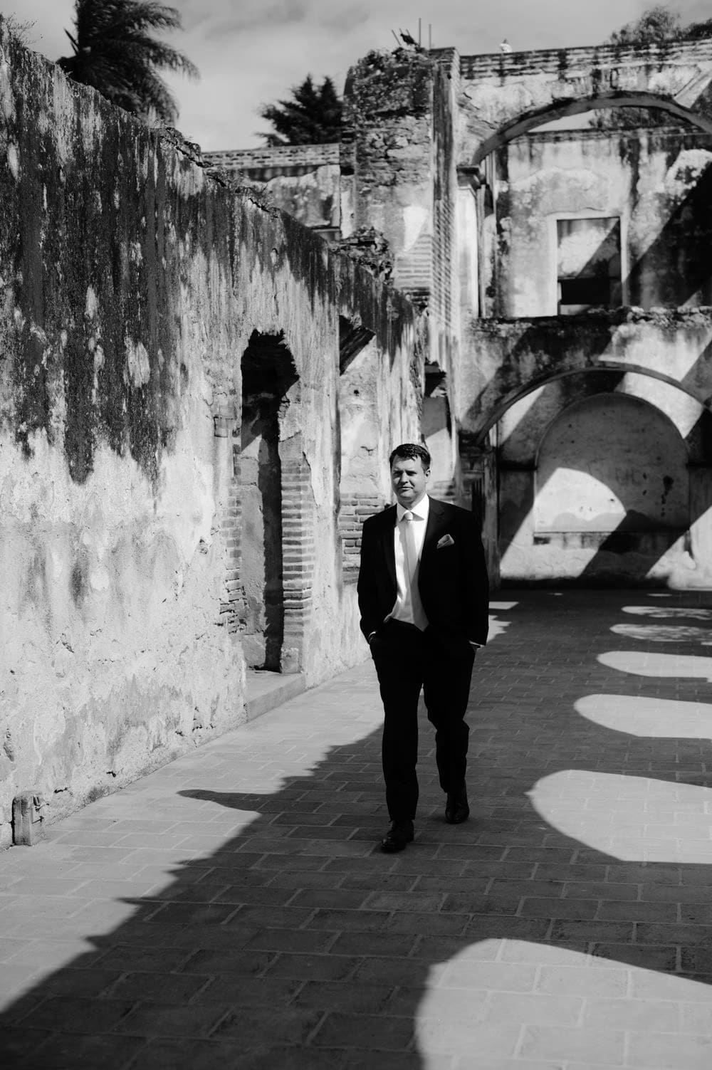 Groom Nathan walks the colonnade of Santa Clara Ruins in black and white — editorial groom portrait in colonial Antigua.