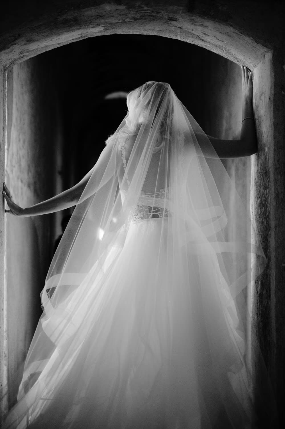 Bride from behind, veil illuminated, framed by a stone arch at Santa Clara Ruins — black-and-white editorial portrait.