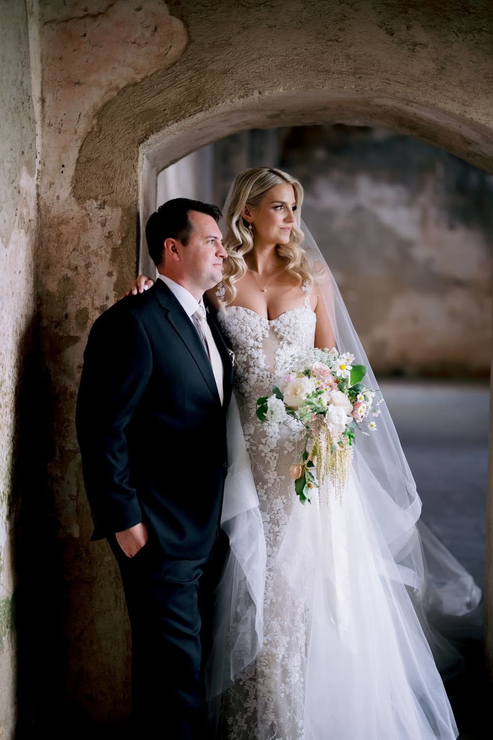 Bride leans on her groom beneath a low arch of Santa Clara Ruins, holding her wildflower bouquet.