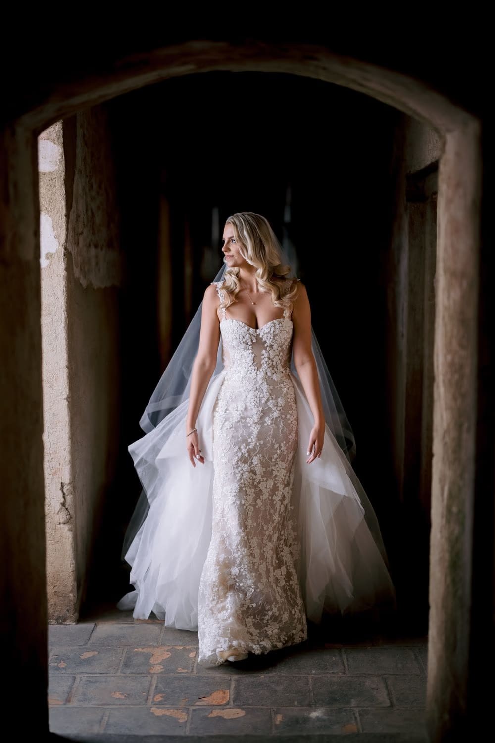 Bride walks through a dim Santa Clara passageway, lace gown catching the side light.