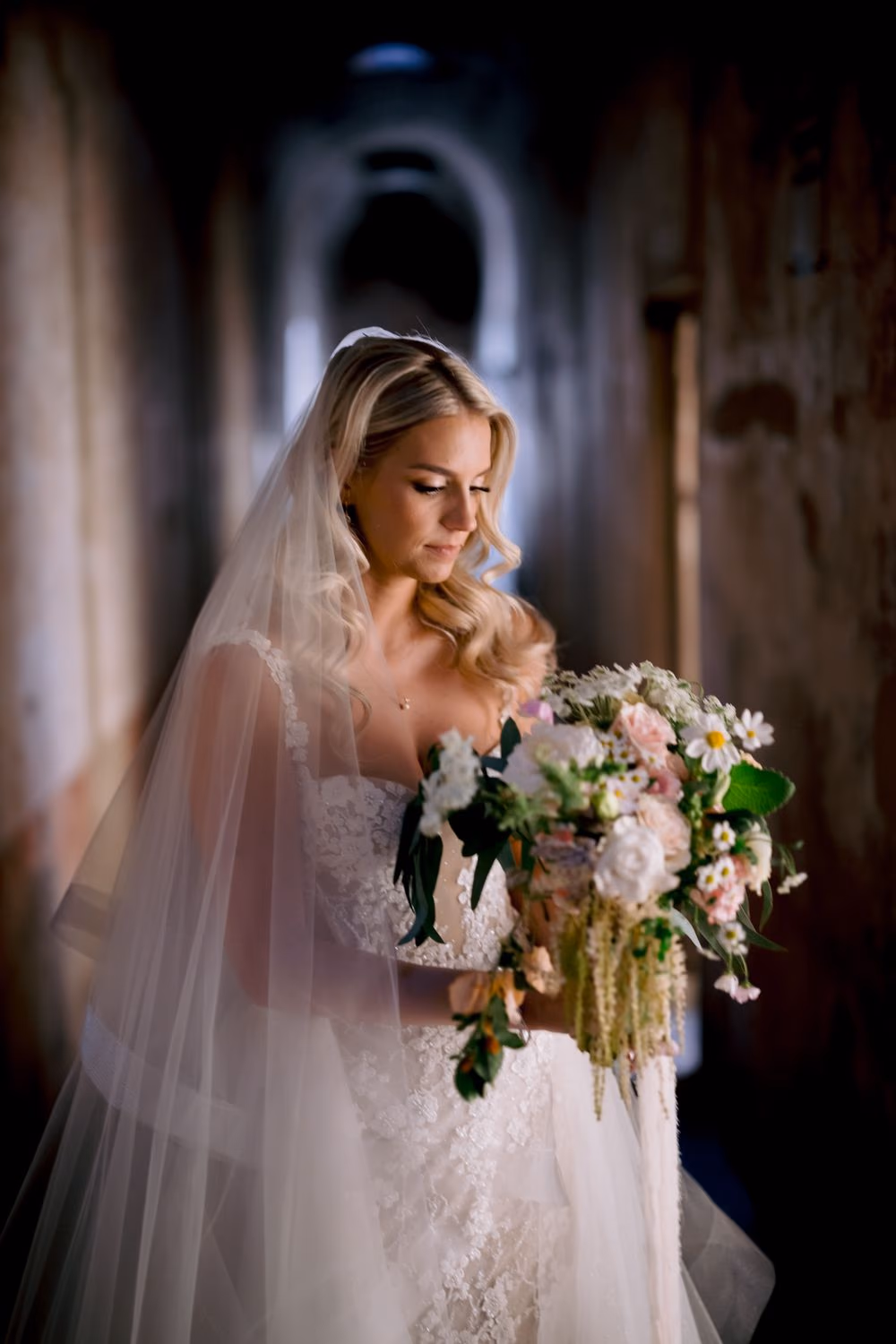 Bride looks down at her wildflower bouquet in a softly lit Santa Clara corridor.