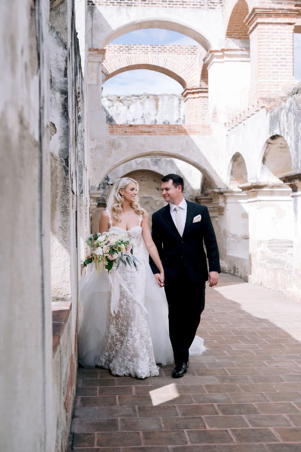 Bride and groom walk a sunlit corridor beneath the brick archways of La Recolección Ruins.