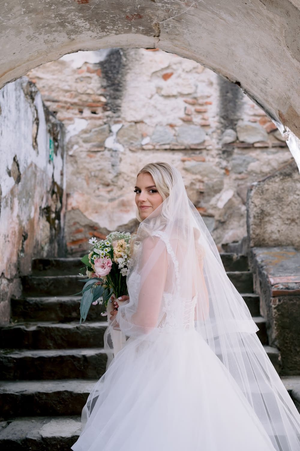 Bride pauses on the rough stone steps of La Recolección Ruins, veil trailing behind her.