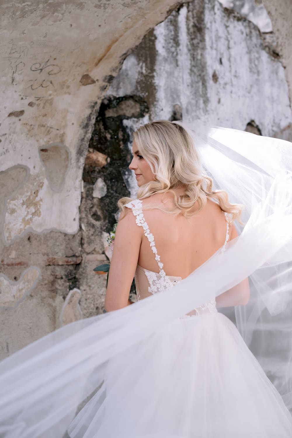 Bride from behind with her veil flowing across a moss-stained stone wall at La Recolección.