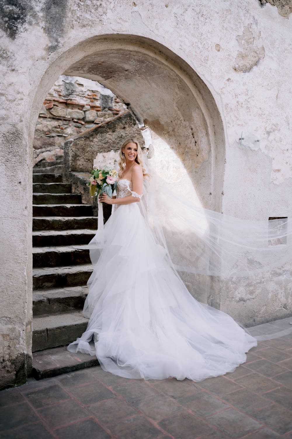 Bride smiles beneath the worn arch of La Recolección's stone staircase with her veil flying out behind her.
