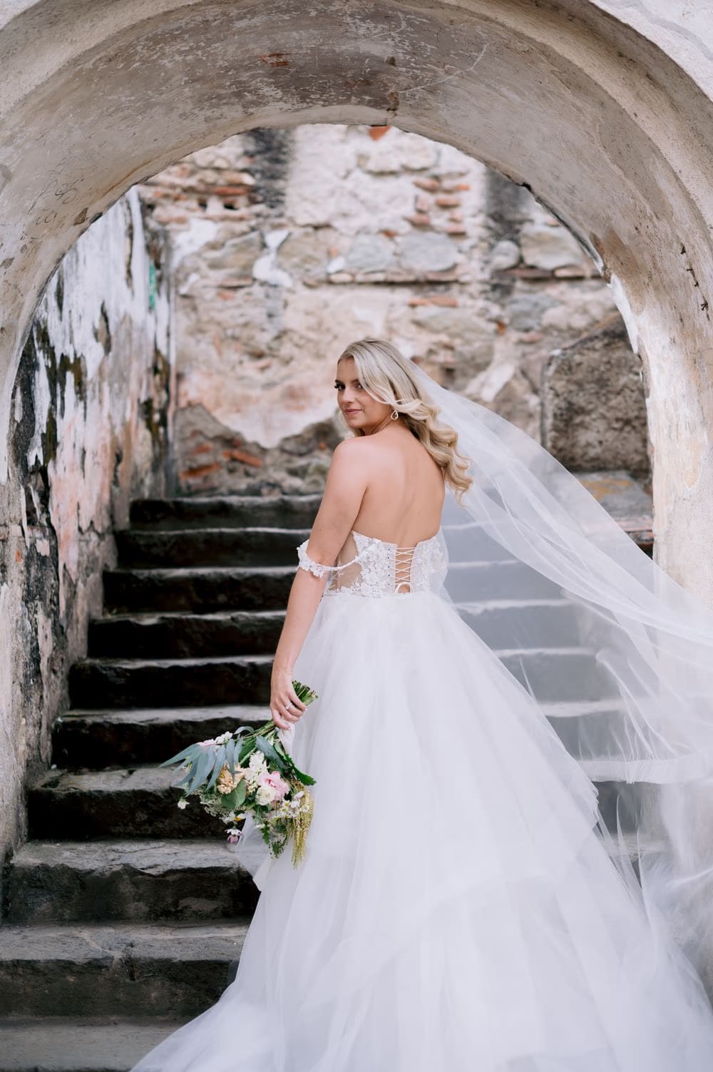 Bride looks back from the stone steps of La Recolección, veil draping into a long train.